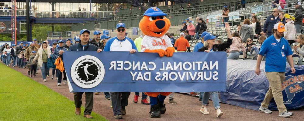 Mascotts, survivors, and others, marching with banner for National Cancer Survivors Day at the stadium